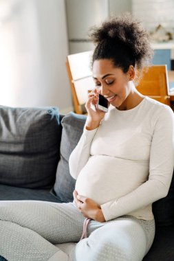 Young beautiful smiling happy pregnant afro woman sitting on sofa holding her belly, looking on it and talking on phone in cozy room at home