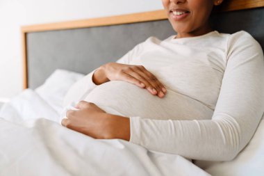 Adult beautiful pregnant smiling african woman holding her belly while liying on bed at home