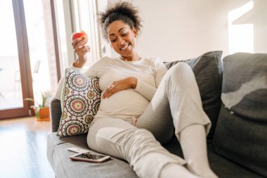 Adult beautiful pregnant smiling african woman eating apple and holding her belly while sitting on sofa at home clipart