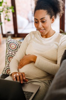 Adult beautiful pregnant smiling african woman working with laptop and holding her belly while sitting on sofa at home