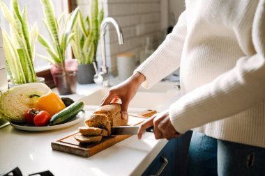 Female hands cutting bread on wooden board next to plate with vegetables closeup