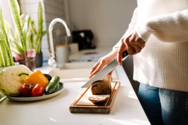 Female hands cutting bread on wooden board next to plate with vegetables closeup