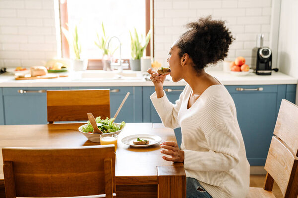 Side view of african woman sitting by table and eating salad with glass of orange juice in cozy kitchen at home