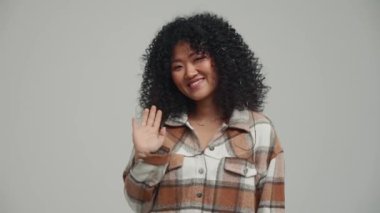 Smiling brunette Asian woman waving hand at the camera in the grey studio