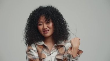 Pretty brunette Asian woman twirling her hair around finger at the camera in the grey studio