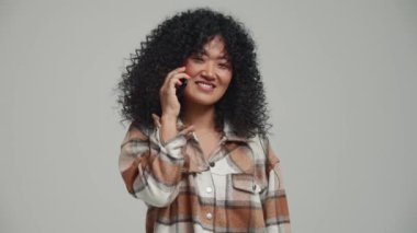 Happy brunette Asian woman talking on mobile in the grey studio