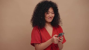 Laughing curly haired Asian woman typing on phone in the beige studio