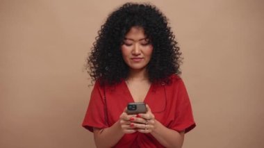 Friendly curly haired Asian woman texting on phone in the beige studio