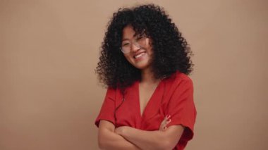 Cheerful curly haired Asian woman wearing eyeglasses looking at the camera in the beige studio