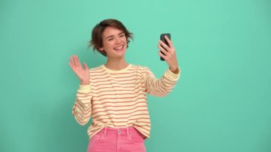 Cheerful young woman with short hairstyle talking by video call on phone in the turquoise studio