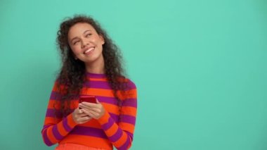 Funny young curly haired woman texting on smartphone in the turquoise studio