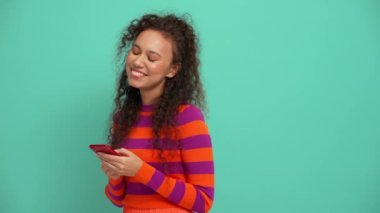 Handsome young curly haired woman texting on smartphone in the turquoise studio