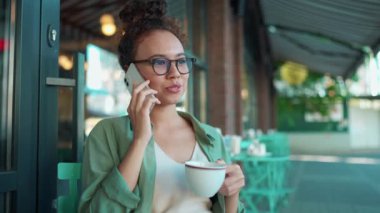Kahkahalar atan, gözlüklü, telefonla konuşan, açık havada kahve içen Afrikalı bir kadın.