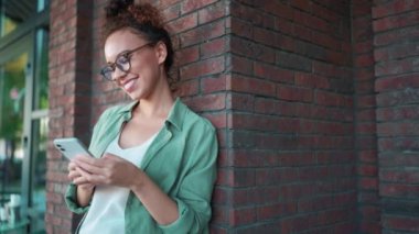 Yeşil tişörtlü ve gözlüklü gülümseyen Afrikalı kadın dışarıda duvarın yanında telefonla yazı yazıyor.