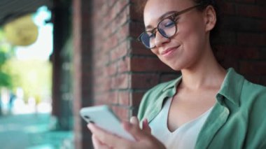 Yeşil tişörtlü ve gözlüklü arkadaş canlısı Afrikalı kadın açık kafede akıllı telefondan yazı yazıyor.