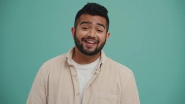 Pozitif Orta Doğulu genç adam kamerayla konuşuyor ve turkuaz stüdyoda bir şeye katılıyor.