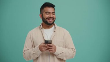 Orta Doğulu gülen genç adam turkuaz stüdyoda cep telefonundan mesaj atıyor.