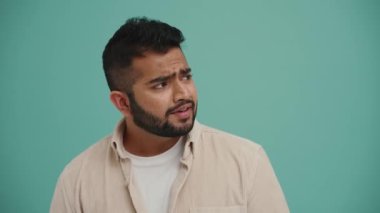 Dalgın Orta Doğulu genç adam kamerayla konuşuyor ve turkuaz stüdyonun yan tarafını işaret ediyor.