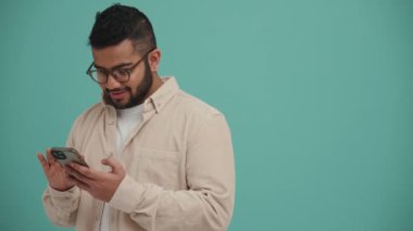 Gözlüklü gülümseyen Orta Doğulu genç adam turkuaz stüdyoda telefonla yazı yazıyor.