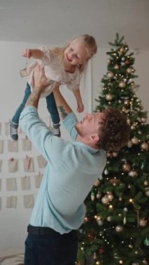 Güzel sarışın baba evde Noel ağacının yanında kızını kollarında döndürüyor.