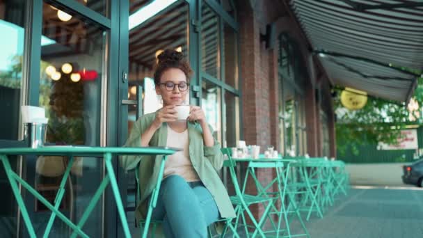 非洲妇女穿着绿色衬衫 戴着眼镜 在户外咖啡馆喝咖啡 获得了放松 — 图库视频影像