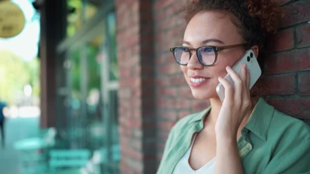 快乐的非洲女人穿着绿色衬衫 戴着眼镜 在户外咖啡店附近用智能手机聊天 — 图库视频影像