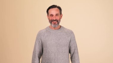 Gri kazak giyen neşeli yetişkin sakallı adam bej stüdyodaki kameraya bakıyor.