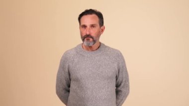 Gri kazaklı, konsantre olmuş yetişkin sakallı adam bej stüdyodaki kameraya sessiz bir jest yapıyor.