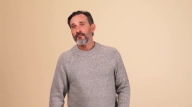 Gri kazak giyen sakallı adam birini arıyor ve bej rengi stüdyoda el sallıyor.