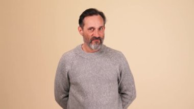 Gri kazaklı kızgın ve sakallı bir adam bej renkli stüdyodaki kameraya bakıyor.