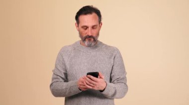 Gri kazaklı, bej renkli stüdyoda cep telefonunda yazan dalgın, sakallı bir adam.