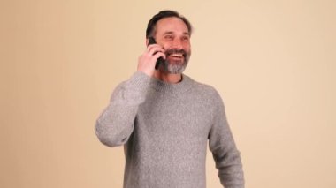 Gri kazaklı, heyecanlı yetişkin sakallı adam bej renkli stüdyoda telefonla konuşuyor.