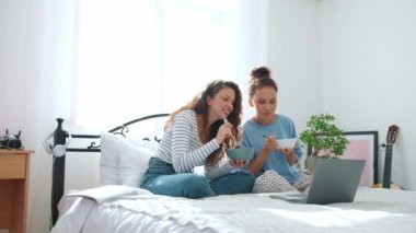Funny girlfriends eating noodles and watching a movie on the bed at home