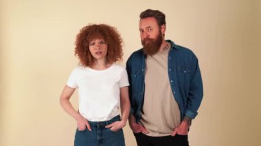 Displeased red haired couple wearing casual clothes showing silence gestures at the camera in beige studio