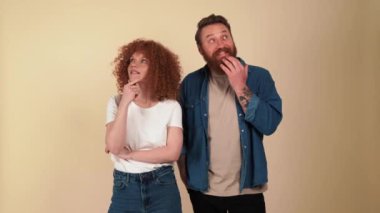 Cheerful red haired couple wearing casual clothes thinking about something in beige studio