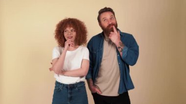 Smiling red haired couple wearing casual clothes thinking about something in beige studio