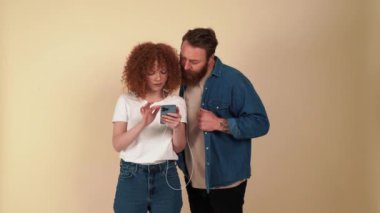 Funny red haired couple wearing casual clothes listening music in headphones on phone in beige studio