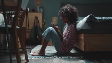 Positive curly haired young African girl typing on phone on the floor near bed at home