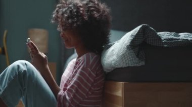 Cheerful curly haired young African girl texting on phone near bed at home