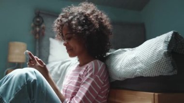 Laughing curly haired young African girl texting on smartphone near bed at home