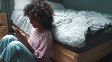 Funny curly haired young African girl typing on smartphone near bed at home