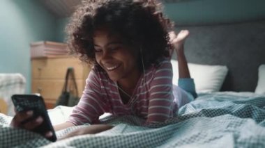 Funny curly haired young African girl talking by video call on phone in headphones on the bed at home