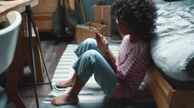 Lovely curly haired young African girl typing on smartphone near bed at home
