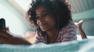 Cheerful curly haired young African girl talking by video call on mobile in headphones on the bed at home
