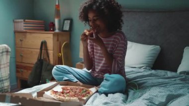 Happy curly haired young African girl eating pizza on the bed at home