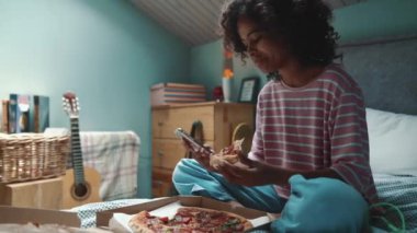 Cute curly haired young African girl eating pizza and typing on mobile on the bed at home