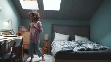 Cheerful curly haired young African girl listening music in headphones and dancing at home