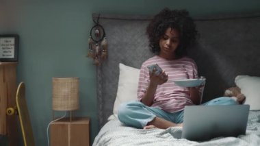 Curly young African girl eats muesli with milk and watches something on the laptop on the bed at home