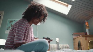 Friendly curly young African girl typing on mobile on the bed at home