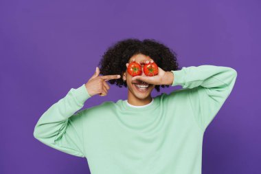 Young man pointing finger at himself while making fun with tomatoes isolated over purple background